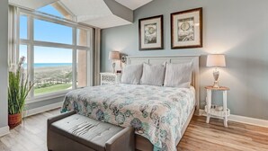 A bedroom with a large window, a bed with a floral bedspread, two bedside tables with lamps, and framed pictures on the wall. - Sailmaker 612 - Hosted By Moore AI Rentals (Fernandina Beach)