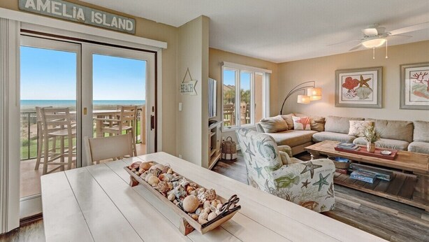 A spacious living area with a view of the ocean, a dining table with a seashell centerpiece, and a sign reading "AMELIA ISLAND". - Sailmaker 612 - Hosted By Moore AI Rentals (Fernandina Beach)