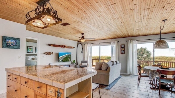 Une cuisine avec un plafond en bois, un grand îlot central doté de tiroirs, un ventilateur de plafond et une vue sur l’extérieur.