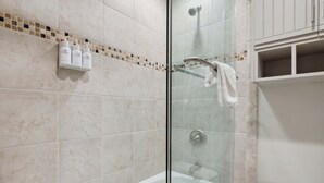 Een moderne badkamer met een douchecabine, een wit bad en een toilet. Boven de douche staan drie flessen shampoo op een plank.