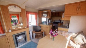 A compact RV interior with a kitchenette, a dining area, a fireplace, and a seating area with cushions. - Beautiful 3-bed Cabin in Pembrokeshire Coast (Haverfordwest)