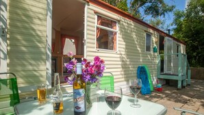 A small, single-story mobile home with a patio area. There is a table with a bottle of beer, a bottle of wine, and a glass of wine. A vase with purple flowers is on the table. - Beautiful 3-bed Cabin in Pembrokeshire Coast (Haverfordwest)