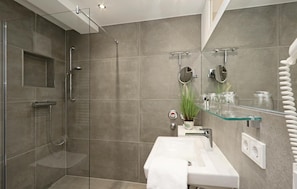A modern bathroom with a glass shower enclosure, a white sink, and a glass shelf with toiletries. - Angel (Binz)