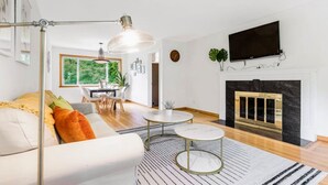 A modern living room with a sofa, coffee table, and fireplace. There is a dining area with a table and chairs, and a large window with a view of greenery.