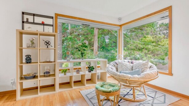 A room with a large window, a wicker chair, a white shelving unit, and a small stool.