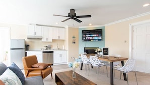 Un salon moderne avec un canapé, une table basse et un coin repas avec des chaises. On y trouve une cuisine avec des meubles blancs et des appareils électroménagers. Un ventilateur de plafond est visible, et un téléviseur à écran plat est fixé au mur.