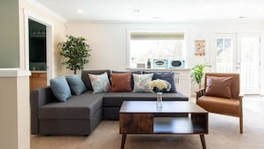 Un salon avec un canapé d’angle, une table basse et un fauteuil. On aperçoit, au fond, une plante, un vase avec des fleurs et un four à micro-ondes.