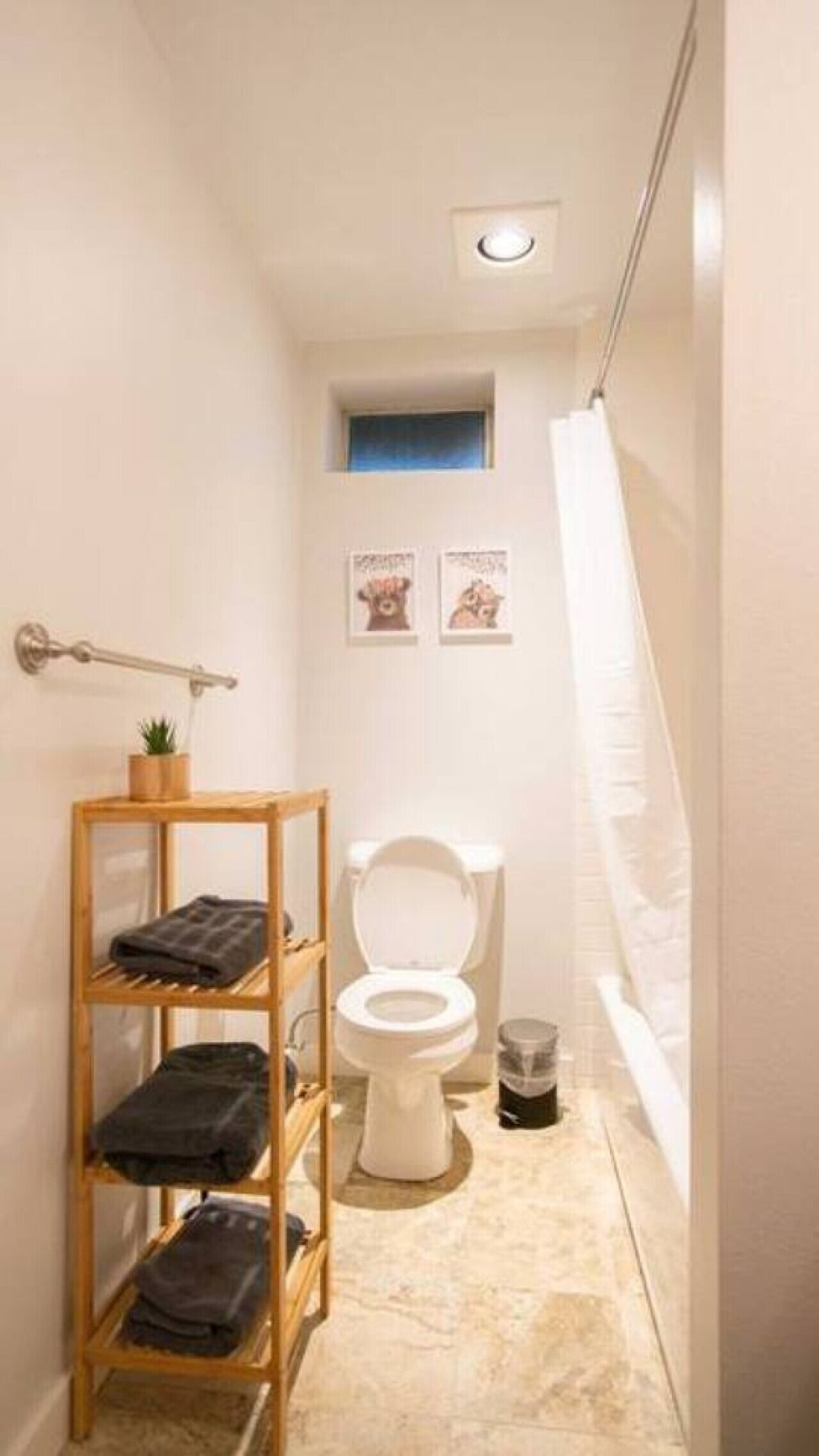 Ein kleines, schmales Badezimmer mit Toilette, Ablage für Handtücher, kleinem Fenster und Duschvorhang.
