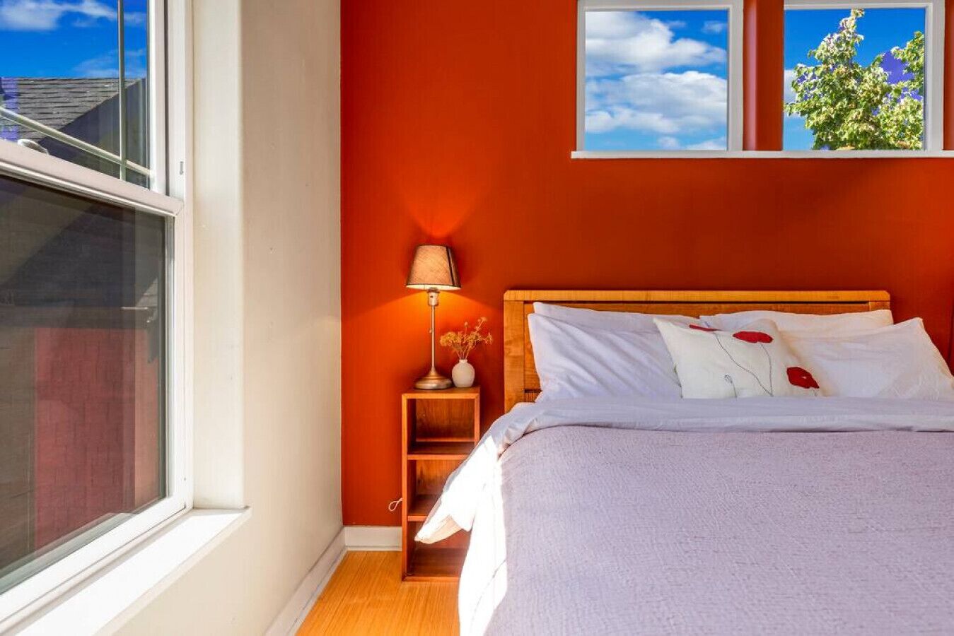 A bedroom with a wooden headboard, a bed with white bedding, a nightstand with a lamp, and a window with a view of the sky and trees.