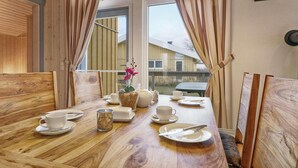 A dining area with a wooden table set for two, a vase with flowers, and a view of a house through large windows. - Amazing Home in Rechlin With 2 Bedrooms, Sauna and Wifi (Rechlin)