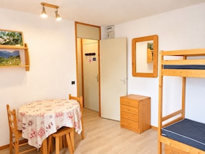 Ein Zimmer mit einem Etagenbett, einem hölzernen Schminktisch, einem Tisch mit gemustertem Tischläufer, einer an der Wand befestigten Ablage mit einem Bild und einer Tür zu einem anderen Raum.