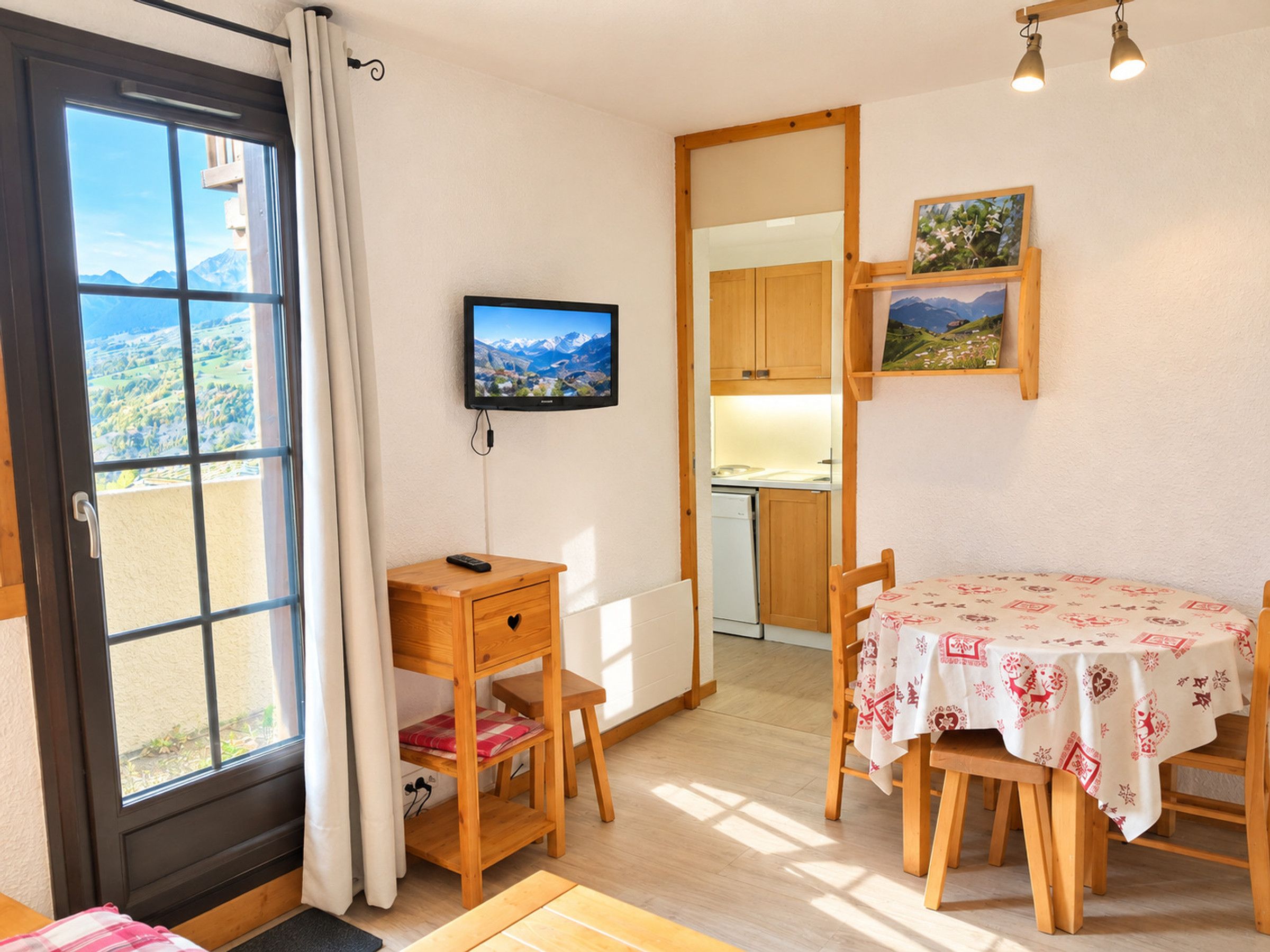 A room with a dining table, a TV mounted on the wall, a kitchen area, and a view through a window.