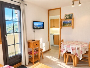 A room with a dining table, a TV mounted on the wall, a kitchen area, and a view through a window. - Cozy central studio, balcony, near slopes (La Plagne Montchavin Les Coches)
