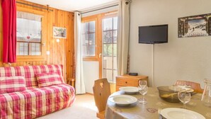 Una habitación acogedora con un sofá de cuadros, una mesa de comedor para dos personas y un televisor montado en la pared.