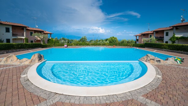 A swimming pool with a clear blue center and a brick border, surrounded by two-story buildings with balconies and satellite dishes. - Appartamento Katy (Polpenazze del Garda)