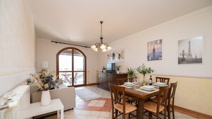 A dining area with a wooden table set for four, surrounded by chairs. There is a white bench with a vase of flowers, a radiator, and a view of the outdoors through a glass door. - Appartamento Katy (Polpenazze del Garda)