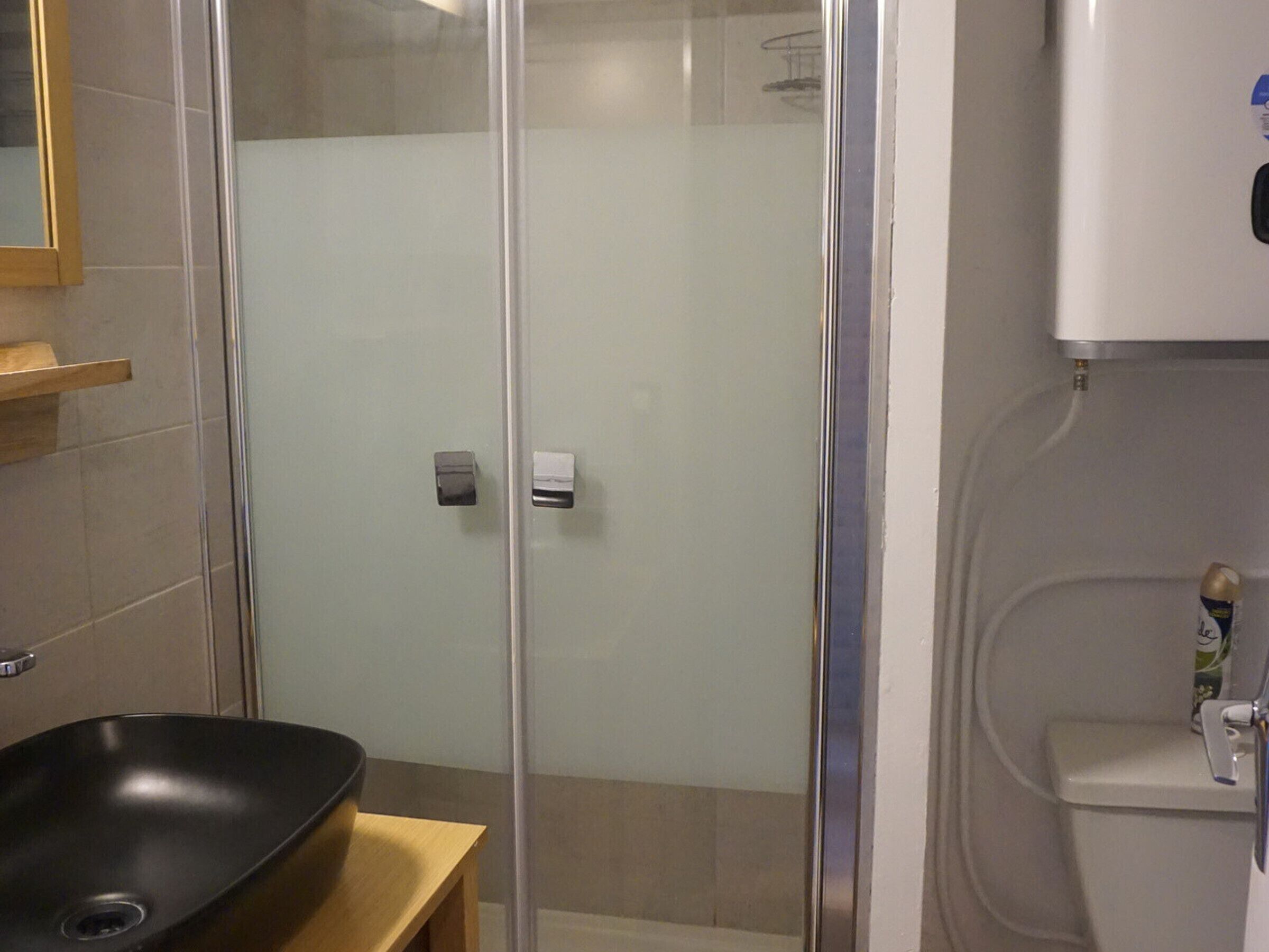 Un baño con ducha envuelta en vidrio, un lavamanos negro y un inodoro.