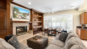 A spacious living room with a fireplace, a large flat-screen TV, a dining area with a table and chairs, and a kitchen area with wooden cabinets. - It's A Mirage - Victoria Falls Home (Rancho Mirage)