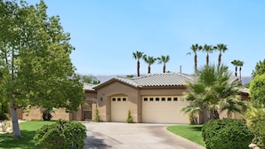 A single-story house with a beige facade, a tiled roof, and a garage. There are palm trees and a well-maintained lawn in front. - It's A Mirage - Victoria Falls Home (Rancho Mirage)