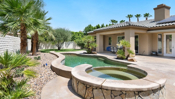 A swimming pool with a stone border, palm trees, and a house with a tiled roof. - It's A Mirage - Victoria Falls Home (Rancho Mirage)
