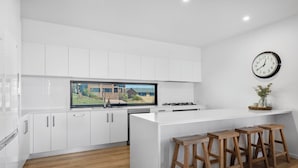 A modern kitchen with white cabinetry, a large window, a gas stove, and wooden stools. - Echoes Luxury Residence, Pet Friendly Walk to Cafes (Woolgoolga)