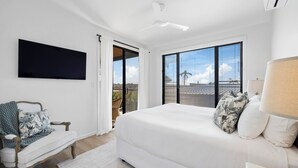 A bedroom with a large bed, a TV mounted on the wall, a chair, and a view of the outdoors through large windows. - Echoes Luxury Residence, Pet Friendly Walk to Cafes (Woolgoolga)