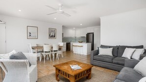 Un soggiorno moderno con un divano grigio, poltrone in vimini bianco, un tavolino in legno e un angolo cottura con pensili bianchi e una ventola a soffitto bianca.