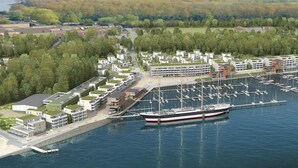 A marina with a large boat docked, surrounded by modern buildings with green roofs, a marina with multiple boats, and a forested area in the background. - Promenadenvilla (Lübeck)