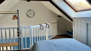 A bedroom with a sloped ceiling, a bed with a patterned cover, a wall clock, a white railing, and a window. - L'hippocampe - Appt Mezzanine Centre D'arromanches (Arromanches-les-Bains)
