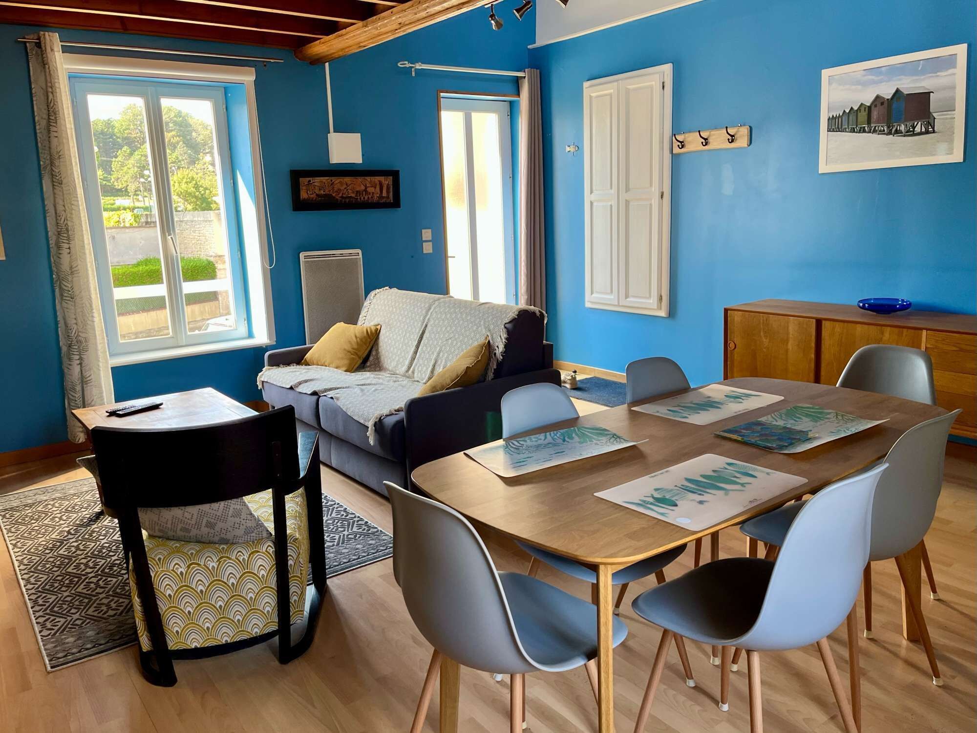 Ein Zimmer mit blauer Wand, einem Sofa, einem Esstisch mit Stühlen, einer kleinen Kommode und einem Fenster mit Vorhängen.
