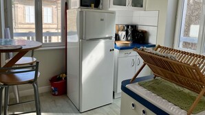 A compact kitchen with a refrigerator, microwave, and a small sink. There is a dining table with chairs and a window providing a view of buildings outside. - L'hippocampe - Appt Mezzanine Centre D'arromanches (Arromanches-les-Bains)