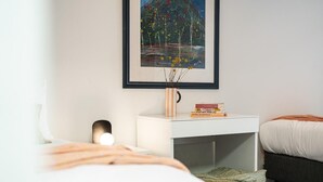Quarto com cama, mesa de cabeceira, abajur, quadro decorativo e pequena estante.