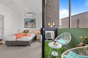 Relax in this serene space with a large bed, bedside table, and framed picture, plus a balcony with a wicker chair and table.