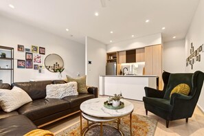 A modern living room with a sofa, armchair, coffee table, and a kitchen area in the background. - StayCentral - Boulevard Gardens (Coburg) - Sacramento Way, Coburg - 3 Bedrooms, 4 Beds, 2 Bathrooms, 2 secure undercover parking spots. (Coburg)