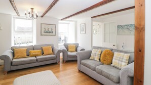 Un salon avec deux canapés gris, chacun agrémenté d’un coussin jaune, un pouf blanc, des poutres en bois au plafond et un lustre.