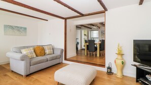 Un salon avec un canapé gris, un pouf à motifs, un plancher en bois, un meuble pour la télévision et un vase rempli de fleurs.