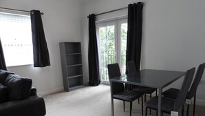 A modern room with a grey sofa, a glass-top dining table with four chairs, a grey shelving unit, and a large window with black curtains. - Beautiful 2-bed Apartment in Poulton-le-fylde (Poulton-le-Fylde)