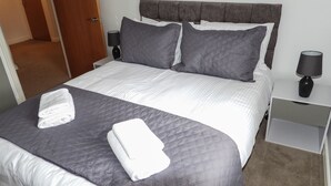 A neatly made bed with a grey quilt, two pillows, and two folded white towels. There is a bedside table with a lamp and a nightstand with a lamp. - Beautiful 2-bed Apartment in Poulton-le-fylde (Poulton-le-Fylde)