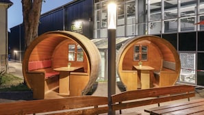 Dos estructuras con forma de barril de madera, con asientos y mesas, ubicadas frente a un edificio moderno con amplios ventanales.