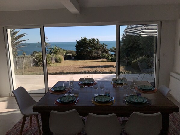Mesa de comedor dispuesta para seis personas, con vista al mar a través de grandes puertas de vidrio.