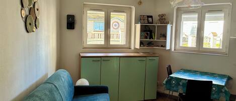 Een kamer met een groene kast, een blauwe bank, een eettafel met een stoel en een raam met uitzicht op gebouwen.