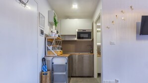 A compact kitchen with stainless steel appliances, a microwave, and a small sink. There is a wooden shelf with kitchen items and a plant. A blue umbrella and a metal cart are visible.