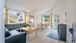 Un vaste espace de vie comprenant un canapé, une table à manger et des chaises. On y trouve également un téléviseur et une cheminée. De grandes fenêtres laissent entrer la lumière naturelle et offrent une vue sur l’extérieur.
