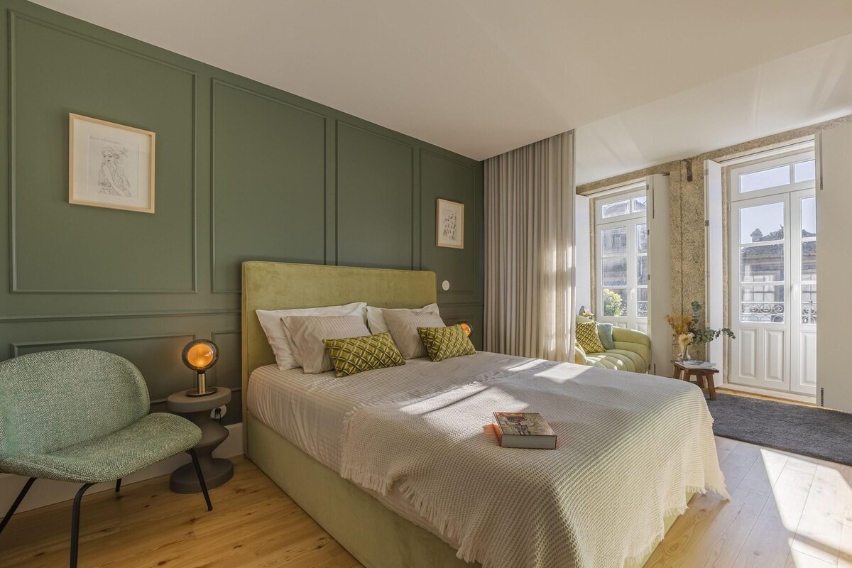 A bedroom with a bed, a chair, a bedside table with a lamp, and a window with curtains.