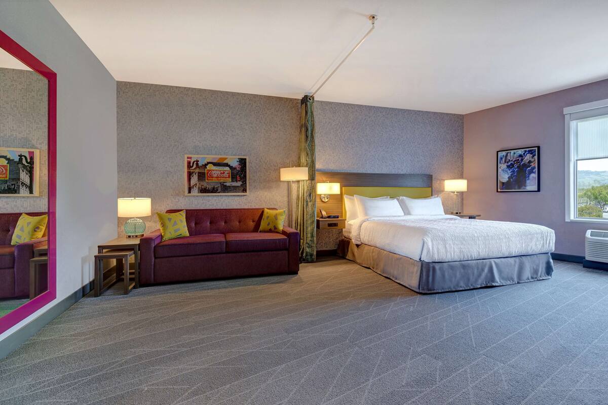 A hotel room with a large bed, a sofa, a desk, and a mirror.