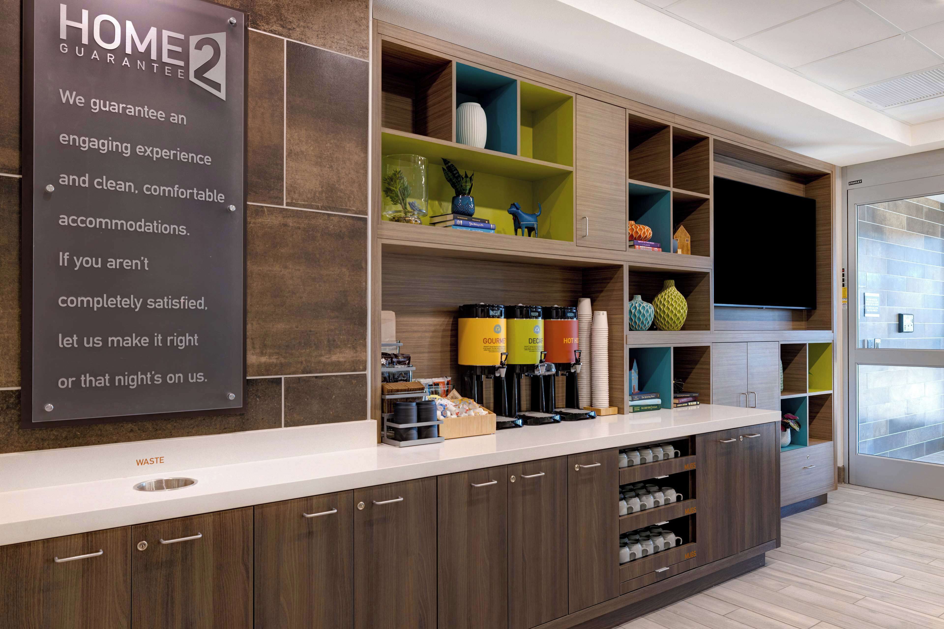 A hotel reception area with a 'Home Guarantee 2' sign, a waste bin, and a counter with coffee machines and a TV.