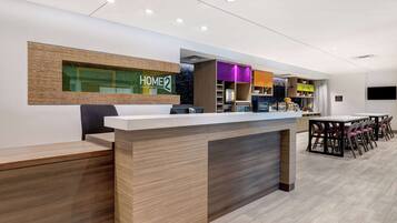 A modern hotel reception area with a wooden reception desk, a digital display, and a seating area with tables and chairs.