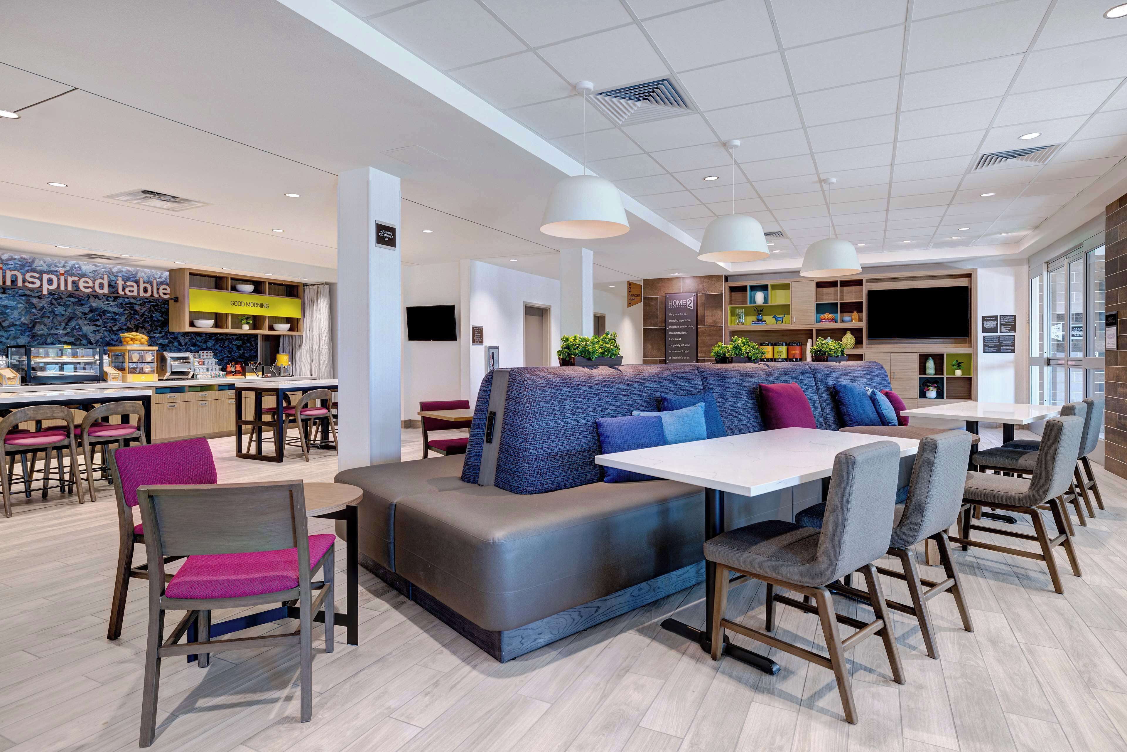 A modern restaurant with a central seating area, a kitchen in the background, and a TV mounted on the wall.