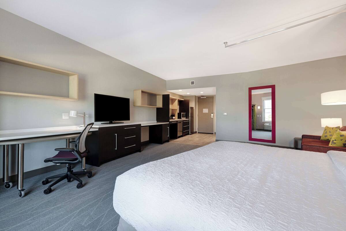 A modern hotel room with a large bed, a desk with a chair, a television, and a mirror.