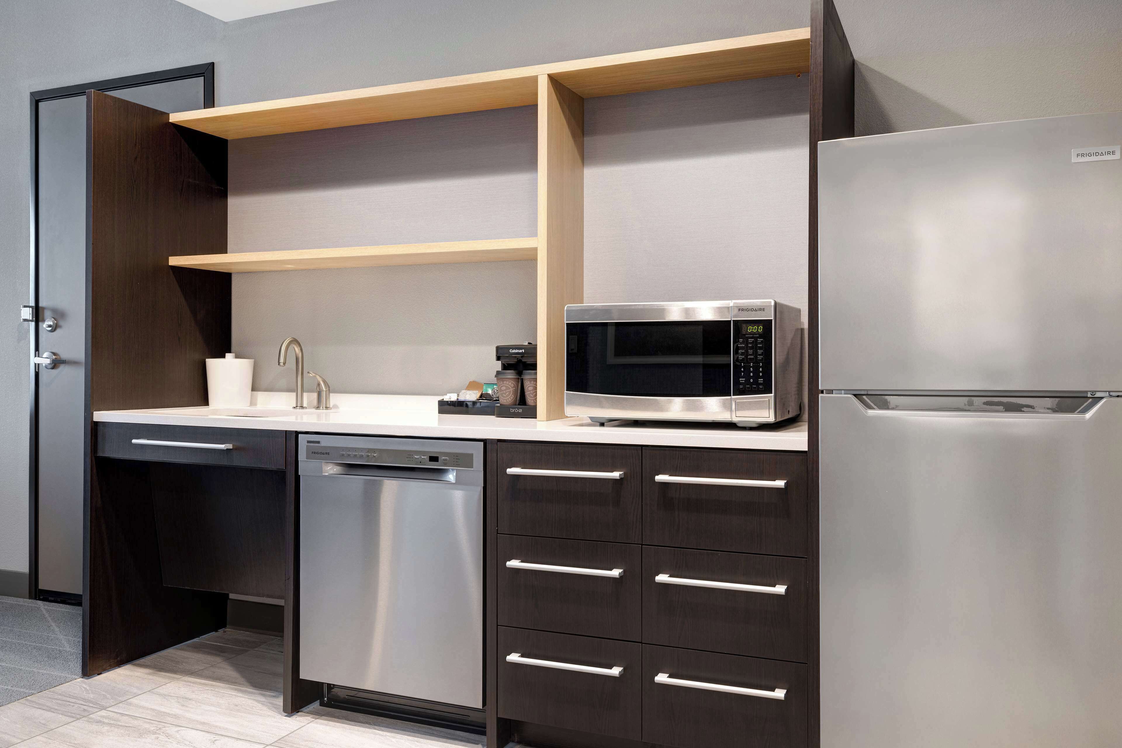 A modern kitchen with a built-in dishwasher, a stainless steel refrigerator, a microwave, and wooden shelves above the countertop.